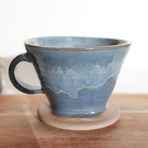 May include: A blue ceramic coffee dripper with a handle, sitting on a beige coaster. The dripper has a light blue glaze with white streaks.