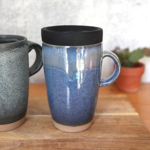 May include: A blue ceramic travel mug with a black lid. The mug has a handle and a speckled, glazed finish. The base of the mug is a light tan color. Another mug is partially visible on the left.