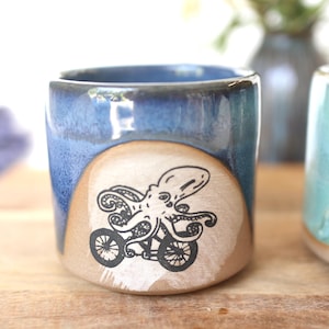 May include: A ceramic mug featuring a blue glazed upper section and a natural tan lower section. A black ink illustration of an octopus riding a bicycle is centered on the tan portion. The mug has a smooth, rounded shape and a glossy finish.