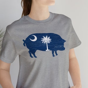 May include: A gray t-shirt with a blue silhouette of a pig. The pig is filled with the South Carolina state flag, which features a crescent moon and a palmetto tree.