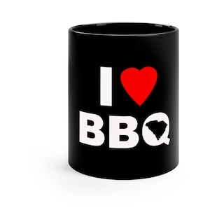 May include: Black coffee mug with white text that says "I love BBQ" with a red heart and a white outline of the state of South Carolina.