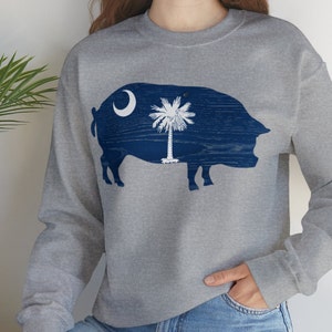 May include: A gray sweatshirt with a blue silhouette of a pig with the South Carolina state flag inside. The flag features a crescent moon and palmetto tree.
