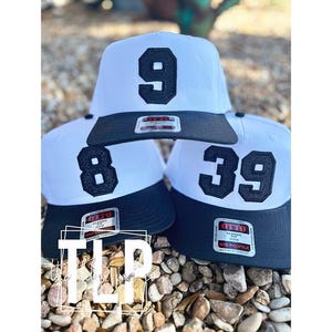 May include: Three white baseball caps with black brims and large black numbers: 9, 8, and 39. Each hat has a classic fit and a mid-profile design. The hats are displayed on a bed of small, light-colored stones.