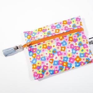 Puede incluir: Un pequeño estuche con estampado floral con una cremallera naranja brillante y una borla azul claro. El estuche está cubierto con un patrón de flores rosas, azules, amarillas y naranjas. Una etiqueta blanca con el texto "HOP LA!" está adjunta.