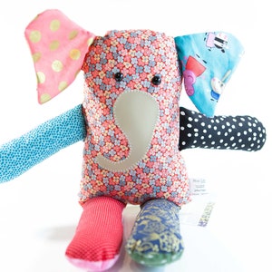 May include: A colorful stuffed elephant toy with a floral patterned body, pink polka dot ears, and blue and black patterned limbs. The elephant has a gray trunk and black button eyes. A tag is attached to the elephant's leg.