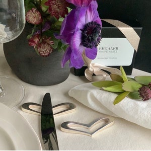 May include: Two silver heart-shaped knife rests on a white linen tablecloth. The knife rests are next to a silver knife and a white napkin. The background includes a vase of purple flowers and a box with the text "Regaler Knife Rests" and the website address "regalerebetsy.com".