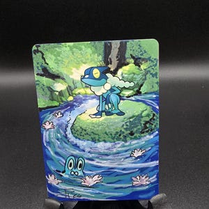 May include: A colorful card featuring a cartoon frog-like creature in a blue and green landscape. The artwork depicts a flowing river with stylized water and floral elements. The card is displayed on a black stand.