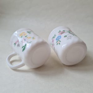 May include: Two white ceramic mugs with a floral pattern in pink, yellow, and blue. The mugs have a slightly textured surface and a handle. The mugs are ideal for serving hot beverages.
