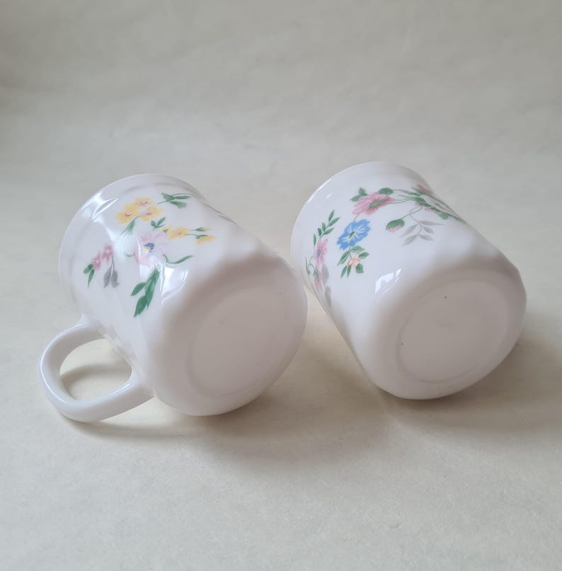 May include: Two white ceramic mugs with a floral pattern in pink, yellow, and blue. The mugs have a slightly textured surface and a handle. The mugs are ideal for serving hot beverages.