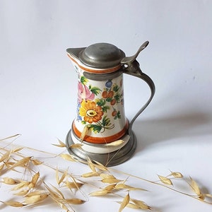 Puede incluir: Una jarra de cerámica vintage con un diseño floral en tonos de rosa, amarillo, naranja y verde. La jarra tiene una tapa y un asa de peltre.