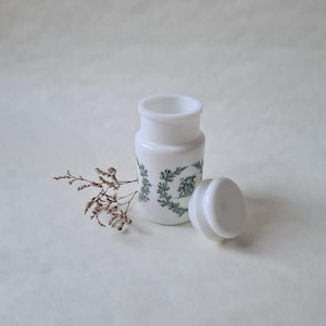 Può includere: Vaso in ceramica bianca con un motivo floreale verde e un coperchio abbinato. Il vaso è vuoto e il coperchio è aperto.