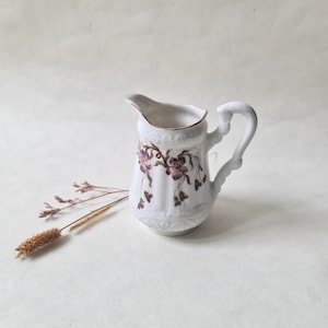 Peut inclure: Un petit pichet en céramique blanche avec une anse et un bec courbés. Le pichet est décoré de motifs floraux violets et d'une bordure dorée. Une brindille d'herbe séchée est à côté du pichet.