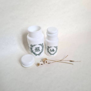 Peut inclure: Deux pots en verre blanc avec des motifs floraux verts et blancs et des couvercles. Les pots sont sur une surface blanche avec des fleurs séchées.
