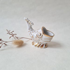 Könnte beinhalten: Ein weißes, fischförmiges Keramikdekorationsstück mit goldenen Akzenten. Der Fisch hat einen gebogenen Schwanz und eine breite Öffnung an der Oberseite. Goldene Details zieren die Flossen, Schuppen und den Sockel. Neutraler Hintergrund.