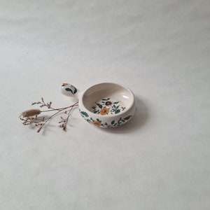 Op de afbeelding: Een klein wit keramisch schaaltje met een handvat. Het schaaltje is versierd met een bloemmotief in tinten geel, groen en bruin.