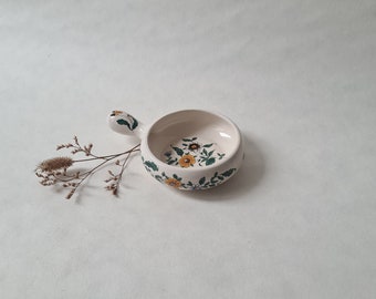 Könnte beinhalten: Eine kleine weiße Keramikschale mit einem Henkel. Die Schale ist mit einem floralen Muster in Gelb-, Grün- und Brauntönen verziert.