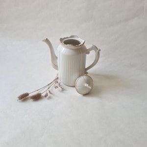 May include: White porcelain teapot with a gold trim and a matching lid. The teapot has a ribbed design and a classic shape.