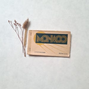 May include: A vintage travel souvenir booklet titled "MONACO" with a gold and teal design. The booklet has the text "10 photographies" and "EDITION S.I.C.A." printed on the cover. Dried flowers are next to the booklet.