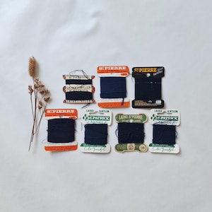 Op de afbeelding: Zeven vintage klossen donker marineblauw stopgaren van wol en nylon van de merken St. Pierre en St. Patrick. Elke klos heeft een uniek papieren label met merktekst en materiaalinformatie. De collectie is gerangschikt met een gedroogde planttak.