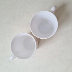 May include: Two white ceramic mugs with handles are displayed against a light background. The mugs are empty, and the interior is visible. The handles are curved and attached to the side of the mugs. The mugs are likely for coffee or tea.