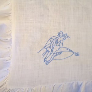 Puede incluir: Una tela de lino blanca con un dise&ntilde;o bordado en azul de dos personas montando una ola.