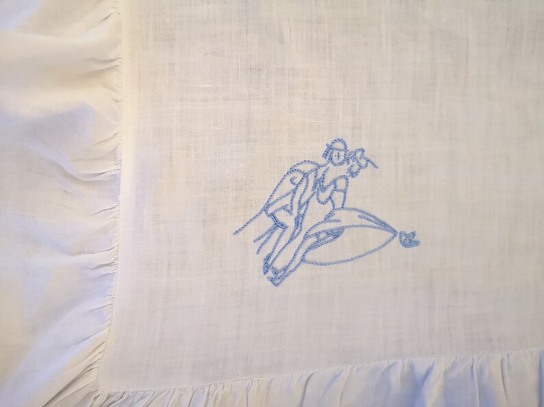 Puede incluir: Una tela de lino blanca con un dise&ntilde;o bordado en azul de dos personas montando una ola.