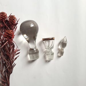 Pode incluir: Três rolhas de garrafa de vidro transparente com designs únicos. Uma é bulbosa e cinza esfumaçado, outra tem uma borda dourada e a terceira tem formato pontiagudo. As rolhas estão dispostas sobre uma superfície branca, com folhagem marrom-avermelhada seca ao fundo.