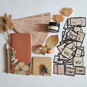 Può includere: Una disposizione piatta di materiali per l'artigianato che include un taccuino marrone con disegni floreali, un ventaglio di legno, una foglia d'oro, nastro washi, un piccolo barattolo di miele e una collezione di adesivi vintage di farfalle.