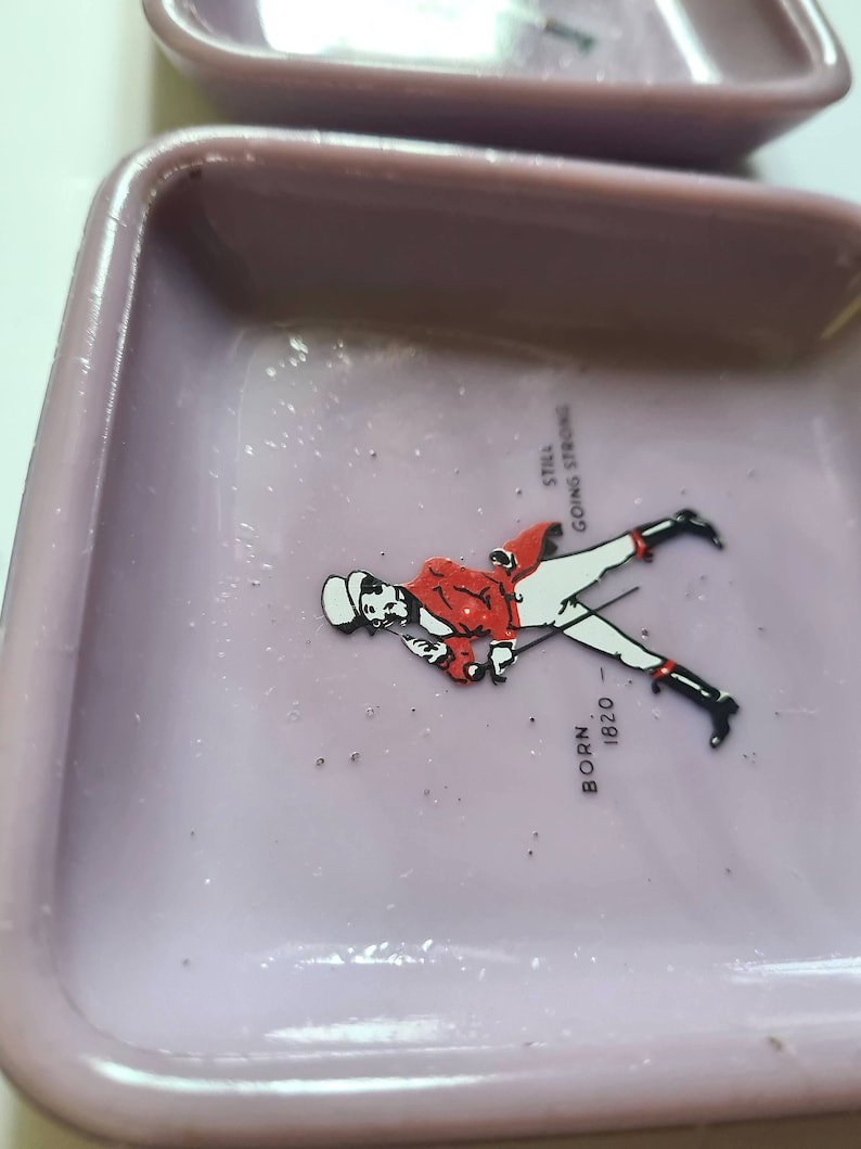 K&ouml;nnte beinhalten: Ein lilafarbener Aschenbecher aus Keramik mit einem Johnnie Walker-Logo, das einen Mann in einer roten Jacke auf einem Pferd zeigt. Das Logo enth&auml;lt auch den Text "Born 1820" und "Still Going Strong".