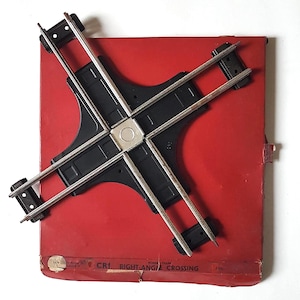 Puede incluir: Un cruce de vías de tren modelo de plástico negro, etiquetado como "CR1 RIGHT-ANGLE CROSSING", sobre un fondo rojo.
