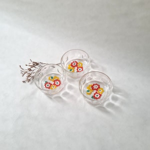 Op de afbeelding: Drie glazen kommen met bloemmotieven. De kommen zijn van helder glas en hebben rode, gele en witte bloemen met groene bladeren.