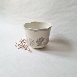 Peut inclure: Un pot en céramique blanc avec un bord festonné et un motif floral avec des fleurs roses et bleues. Le pot est posé sur une surface blanche.