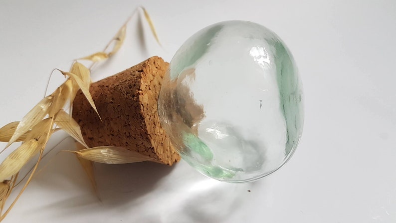 May include: A clear glass bottle stopper with a cork base. The bottle stopper is sitting on a white surface.
