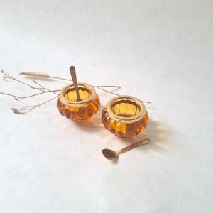Peut inclure: Deux salières en verre ambré avec des bords dorés et des cuillères assorties. Les salières ont un design côtelé. Un brin de fleurs séchées est visible en arrière-plan, sur fond blanc.