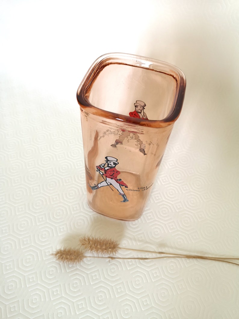 Peut inclure: Un verre &agrave; boire en verre transparent de forme carr&eacute;e avec une teinte rose. Le verre est orn&eacute; d'une image imprim&eacute;e d'un homme en manteau rouge et pantalon blanc, tenant une canne. Le texte "Still Going Strong" est imprim&eacute; sur le verre.