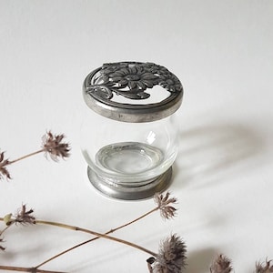 May include: A clear glass jar with a round body and a silver-colored metal rim and base. The lid is also silver-colored and features a floral design. The jar is likely a decorative item or a small vase.