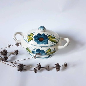 Peut inclure: Un sucrier en céramique blanche avec un couvercle, orné d'un motif floral peint à la main en bleu et vert. Le bol a deux anses et une base ronde.