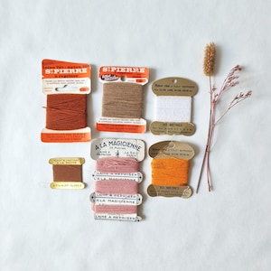 Può includere: Una collezione di cartelle di filato da rammendo vintage in vari colori tra cui ruggine, marrone chiaro, bianco, rosa e arancione, con marchi come St. Pierre e A La Magicienne, accompagnata da un singolo stelo botanico essiccato con piccoli boccioli marroni.