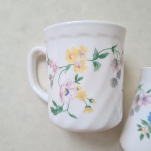 May include: White ceramic mug with a handle, decorated with a floral pattern in yellow, pink, and blue. The flowers are connected by green vines. The mug has a ridged design.