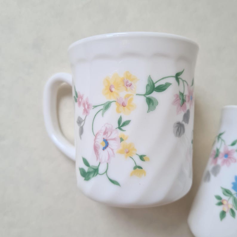 May include: White ceramic mug with a handle, decorated with a floral pattern in yellow, pink, and blue. The flowers are connected by green vines. The mug has a ridged design.