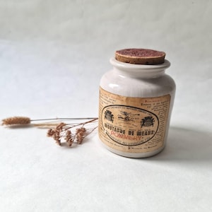 Peut inclure: Un pot en céramique blanc cassé avec un bouchon en liège. Le pot porte une étiquette avec du texte français, dont "MOUTARDE DE MEAUX POMMERY". L'étiquette a un aspect vieilli et usé. Une brindille de fleurs séchées est à côté du pot.
