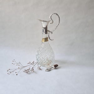 Peut inclure: Une carafe en verre transparent avec un motif en losanges, une poignée et un bec verseur argentés. Un bouchon argenté avec un motif décoratif et un dessus en liège est posé à côté de la carafe. Une brindille de fleurs séchées est au premier plan.