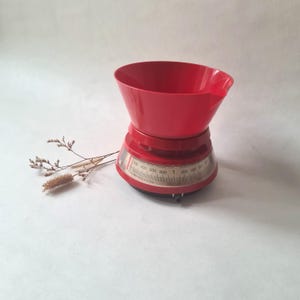 May include: A vintage red kitchen scale with a large bowl and a circular dial. The scale has a capacity of 1 litre. The bowl has a pouring spout. The scale is a classic design, perfect for measuring ingredients.