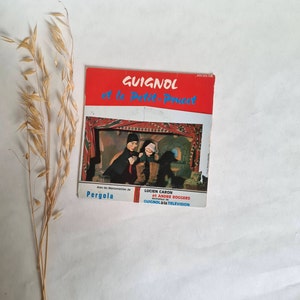 Peut inclure: Une couverture de livre rouge et blanche avec le titre "Guignol et le Petit Poucet" en texte noir. La couverture présente une photo de deux marionnettes, l'une en noir et l'autre en blanc, se produisant sur scène. Le texte "Avec les Marionnettes de Pergola" et "LUCIEN CARON et ANDRE ROGGERS Animateur de GUIGNOL à la TELEVISION" est également sur la couverture.