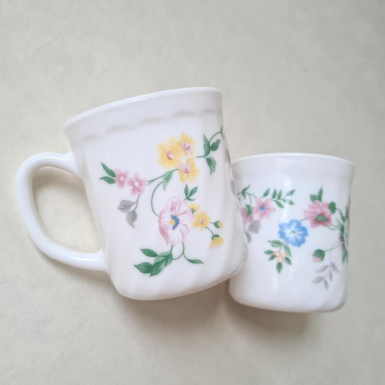 May include: Two white ceramic mugs with floral designs. The larger mug has a handle and a scalloped rim. The floral pattern includes pink, yellow, and blue flowers with green leaves. The smaller mug is cylindrical and also features the floral pattern.