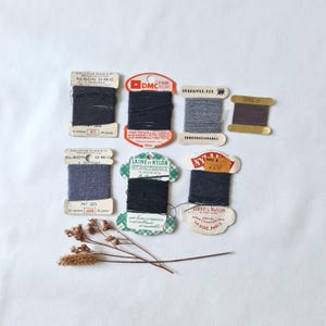 Könnte beinhalten: Eine Sammlung von sechs Vintage-Stopfgarnspulen in Schwarz-, Grau- und Dunkelblautönen mit verschiedenen Papieretiketten von Marken wie DMC und St. Patrick, begleitet von einem kleinen, getrockneten, hellbraunen botanischen Zweig.