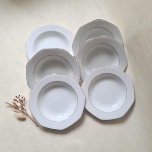 Puede incluir: Un conjunto de siete cuencos de sopa blancos octagonales con un borde dorado. Los cuencos están dispuestos en grupo, mostrando sus líneas limpias y su diseño elegante. Una ramita de flores secas añade un toque de contraste natural.