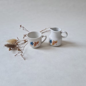 May include: A white teacup and creamer set with a floral design featuring blue and yellow flowers. The set is on a white background.