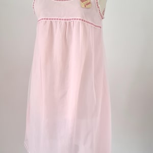 Può includere: Camicia da notte rosa chiaro senza maniche con finiture in pizzo bianco e dettagli rosa. La camicia da notte ha uno scollo tondo e un'etichetta con un'illustrazione di un ventaglio. L'orlo e lo scollo sono rifiniti con pizzo bianco e dettagli rosa.