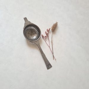 Könnte beinhalten: Ein silberner Vintage-Teesieb mit langem Griff und perforierter Schüssel. Der Sieb liegt auf einer weißen Oberfläche, begleitet von getrockneten Blumen. Der Griff hat ein dekoratives Design, und die Schüssel hat kleine Löcher zum Filtern von Teeblättern.
